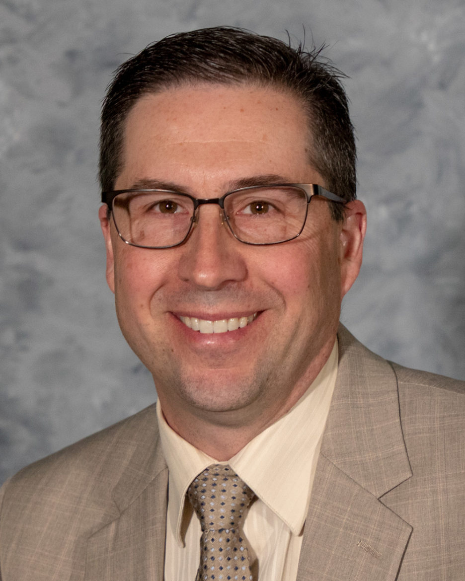 Man with glasses, dark hair and a tan suit smiles. 