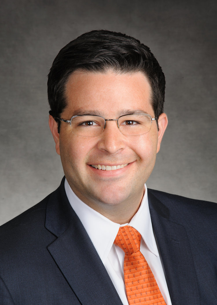 Man with glasses and dark hair, smiling and wearing black jacket, orange tie and white shirt. 